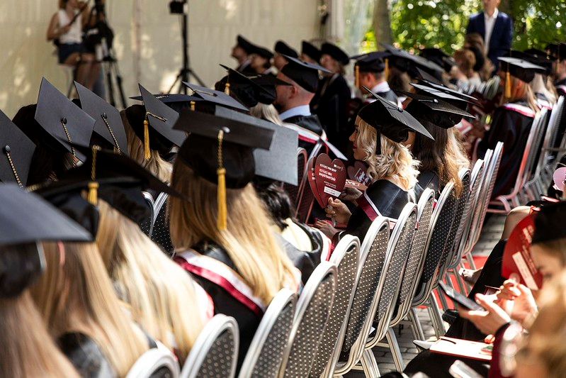RSU absolventi, tērpušies talāros, sēž izlaidumu teltī Iedvesmas parkā