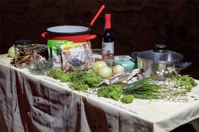 Table displaying cooking ingredients and utensils, including onions, herbs, dried mushrooms, pots and a bottle of wine.